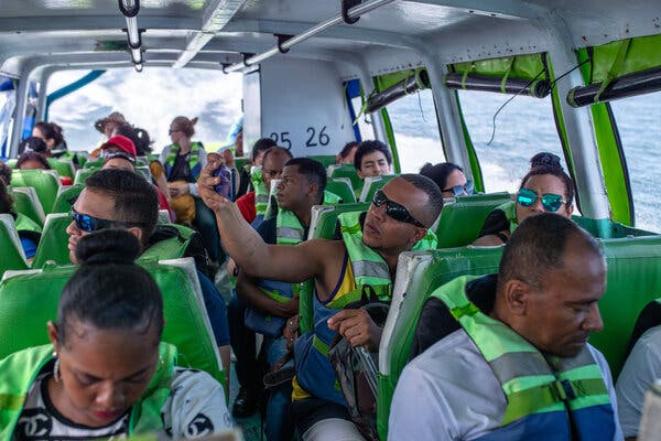 People wearing bright green life jackets sitting on green seats aboard a boat traveling through the ocean. The boat is leaving a broad wake. One man wearing sunglasses is taking a selfie.