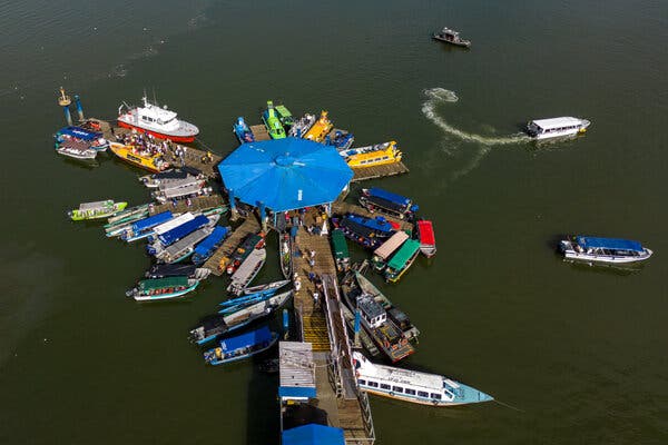 A wooden pier packed with multicolored boats seen from above. The long wooden walkway fans out into several smaller docks at the end. There is an octagonal blue roof over the end of the pier.